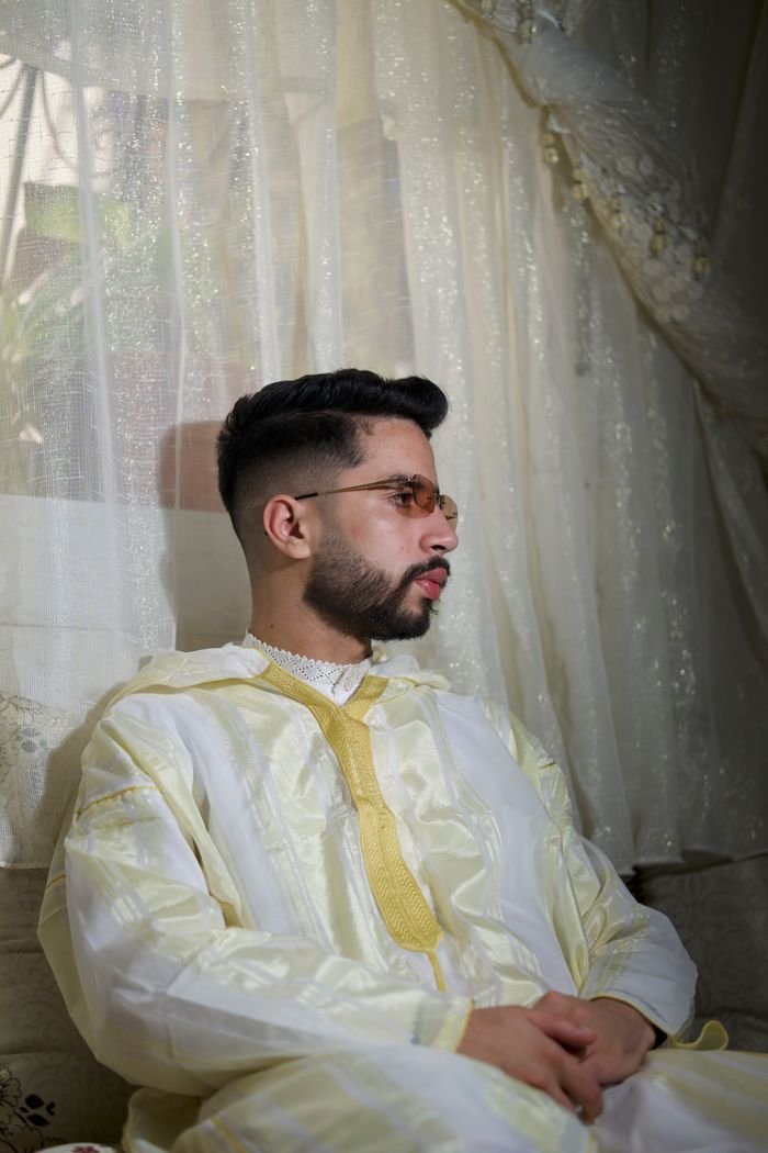 A man in traditional ethnic clothing sitting indoors by a window draped with curtains.